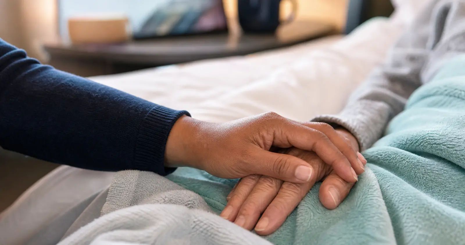 A close view of one person's hand resting gently over a patient's hand on a soft blanket — a quiet moment of reassurance.
